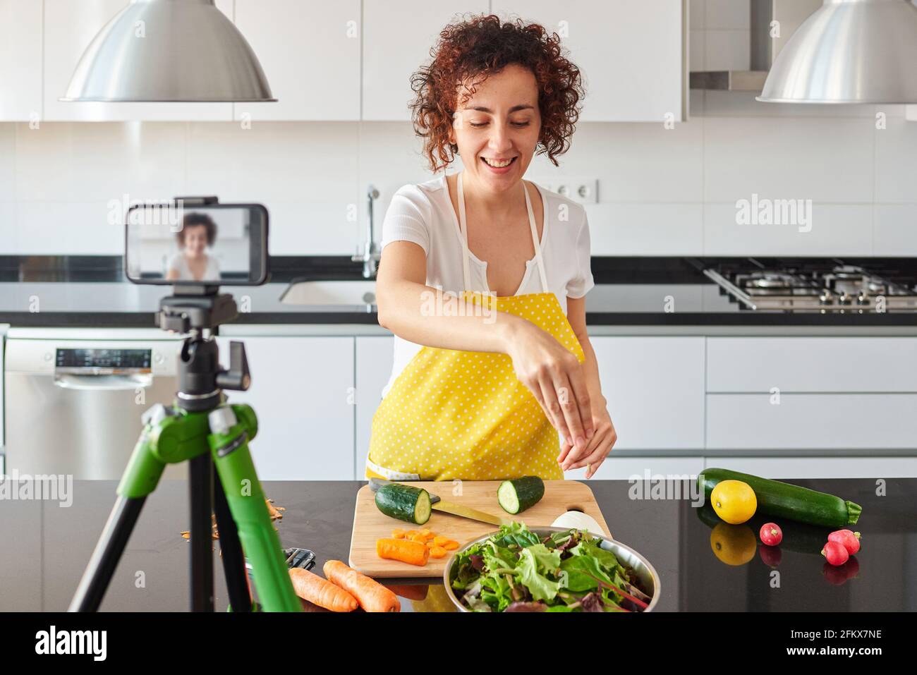 La donna youtuber si registra con il suo smartphone in cucina mentre si prepara una ricetta di insalata, c'è luce naturale e lei indossa una a gialla Foto Stock