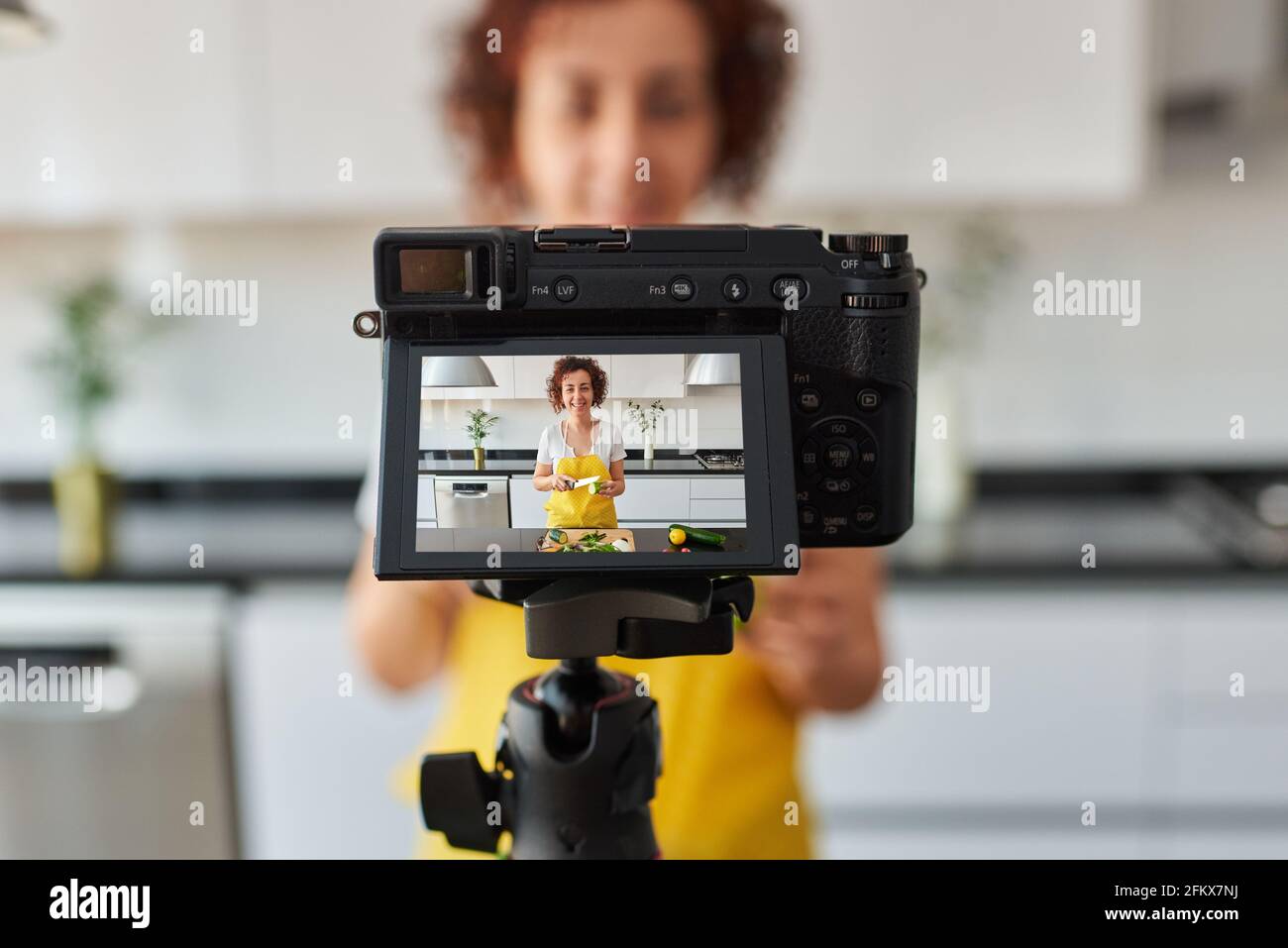 La donna youtuber si registra con una macchina fotografica nella cucina della sua casa mentre prepara una ricetta di insalata, c'è luce naturale e lei indossa un giallo Foto Stock