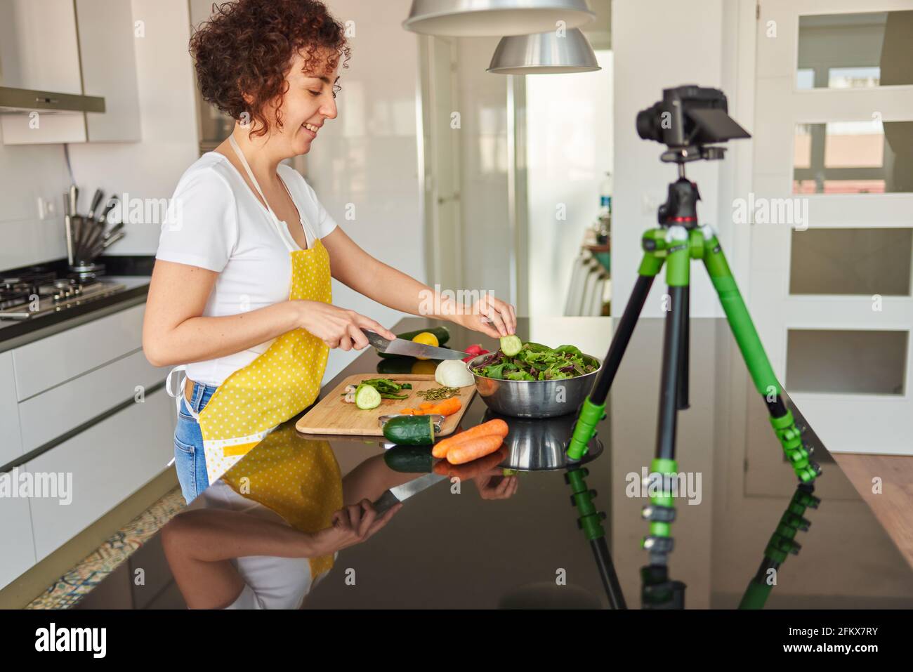 La donna youtuber si registra con una macchina fotografica nella cucina della sua casa mentre prepara una ricetta di insalata, c'è luce naturale e lei indossa un giallo Foto Stock