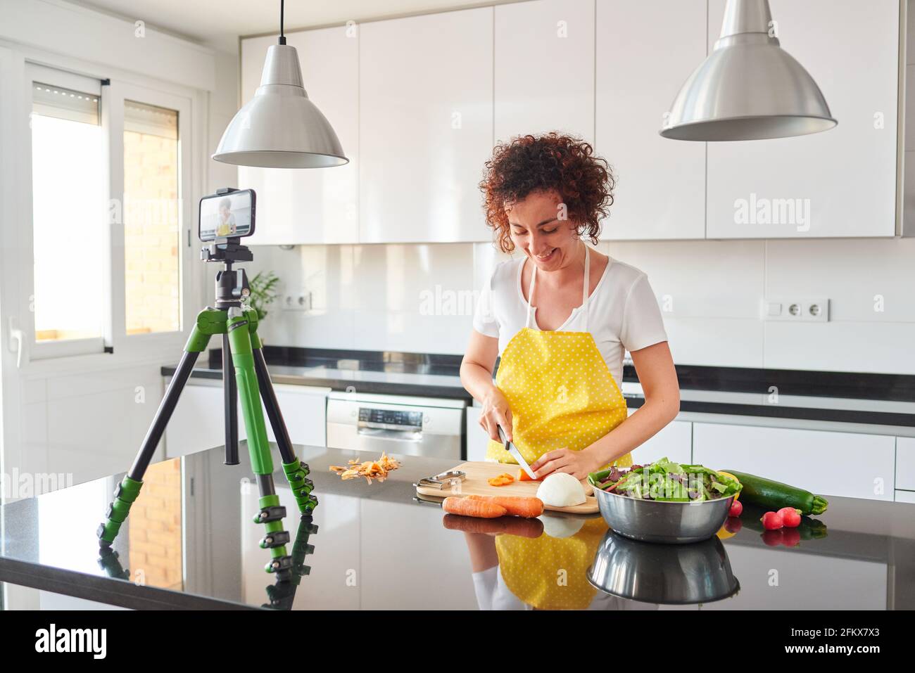 La donna youtuber si registra con il suo smartphone in cucina mentre si prepara una ricetta di insalata, c'è luce naturale e lei indossa una a gialla Foto Stock