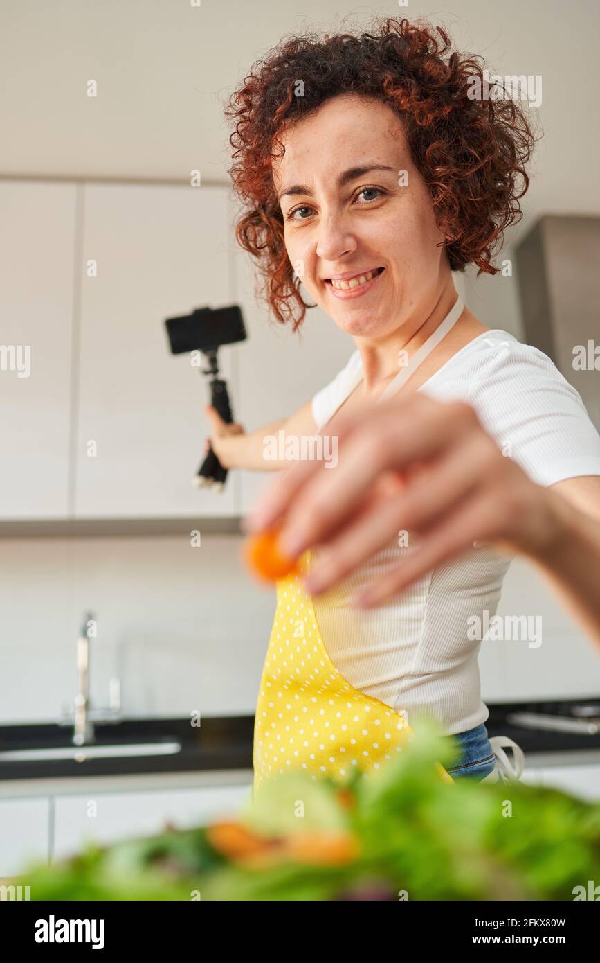 La donna youtuber si registra con il suo smartphone in cucina mentre si prepara una ricetta di insalata, c'è luce naturale e lei indossa una a gialla Foto Stock