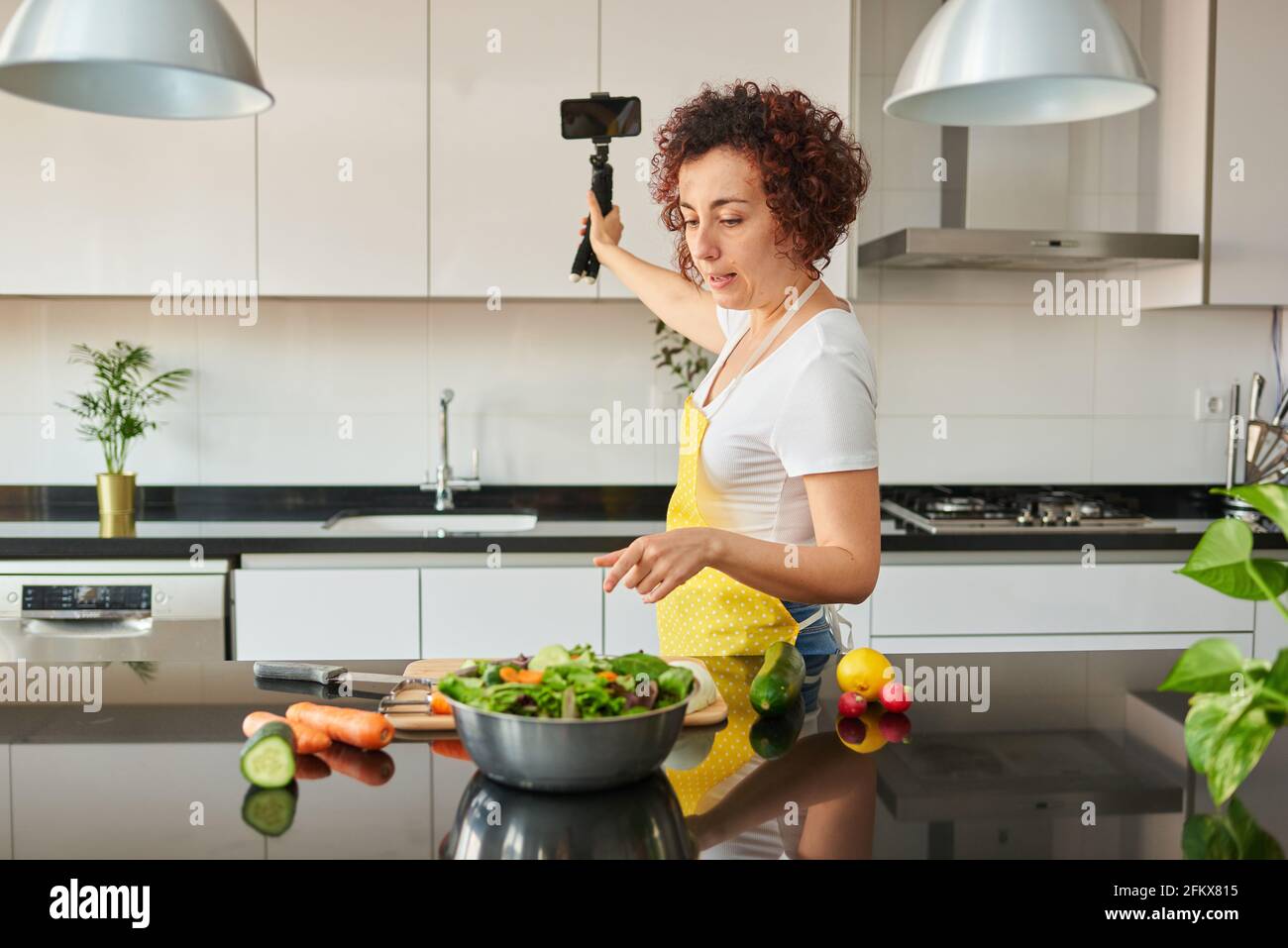 La donna youtuber si registra con il suo smartphone in cucina mentre si prepara una ricetta di insalata, c'è luce naturale e lei indossa una a gialla Foto Stock