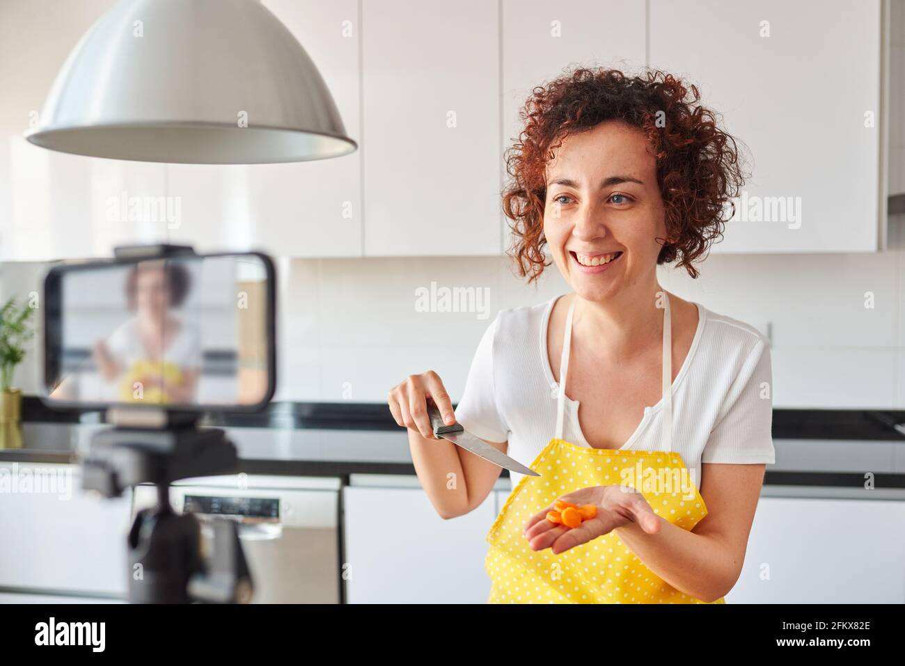 La donna youtuber si registra con il suo smartphone in cucina mentre si prepara una ricetta di insalata, c'è luce naturale e lei indossa una a gialla Foto Stock