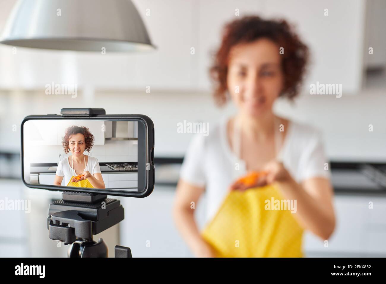 La donna youtuber si registra con il suo smartphone in cucina mentre si prepara una ricetta di insalata, c'è luce naturale e lei indossa una a gialla Foto Stock
