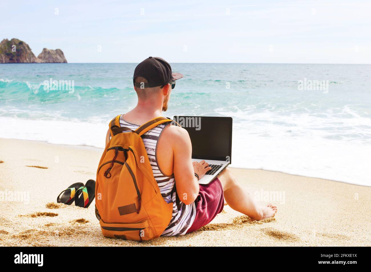 Programmatore di muscoli in forma giovane uomo seduto sulla spiaggia di sabbia, zaino, occhiali da sole, computer portatile, blog di codifica, navigazione. Freelance, con lavoro a distanza Foto Stock