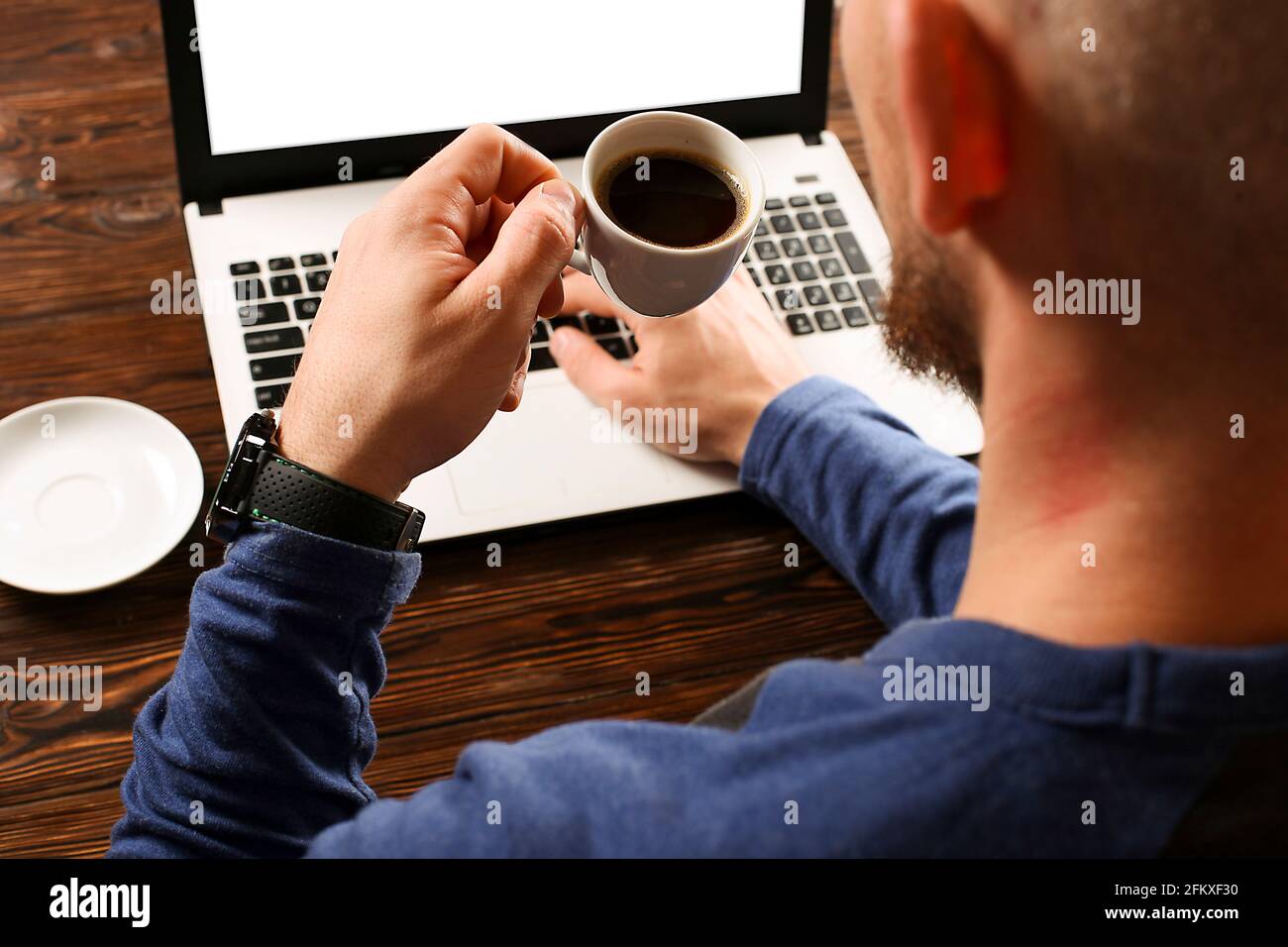 Uomo blogger bearded seduto al tavolo testurizzato in legno, tenendo una tazza di caffè nero, navigare, scrivere sul suo computer portatile. Mani maschili che surfing l'int Foto Stock