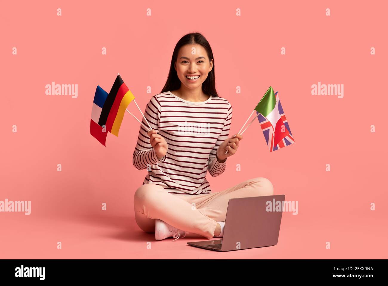 Imparare le lingue online. Happy Asian Woman che tiene bandiere internazionali e che usa il laptop Foto Stock