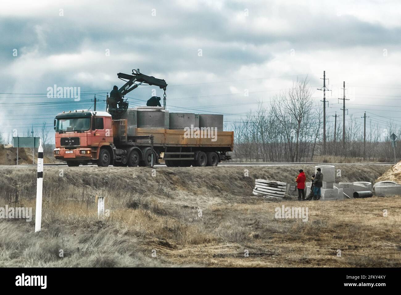 Bielorussia, regione di Minsk - 11 dicembre 2019: I lavoratori edili scaricano o caricano strutture ad anello in calcestruzzo da un autocarro o da un veicolo in un'area industriale. Foto Stock