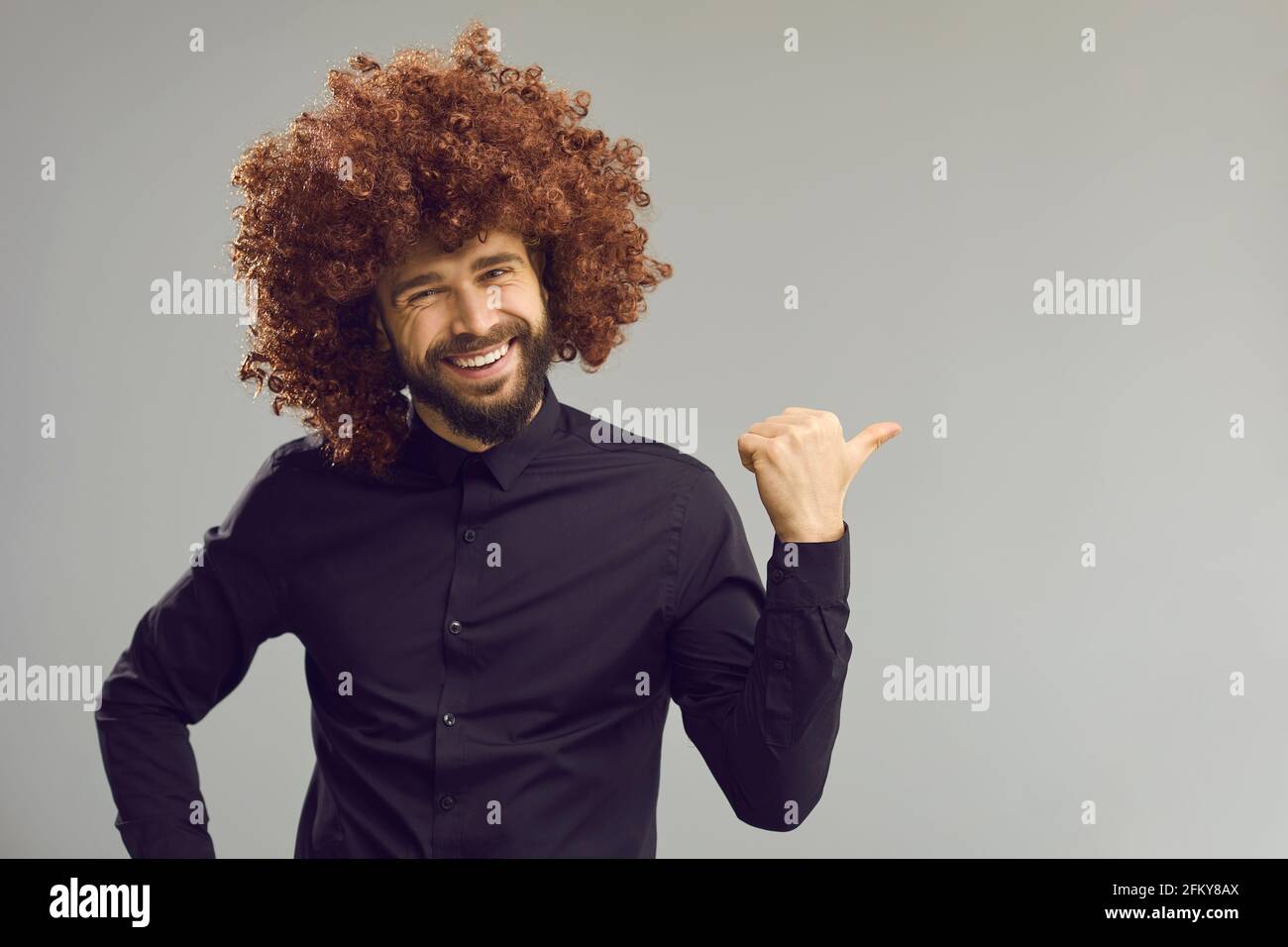 Uomo divertente in parrucca arricciata che punta il pollice da parte a vuoto sfondo dello spazio di copia pubblicitario Foto Stock