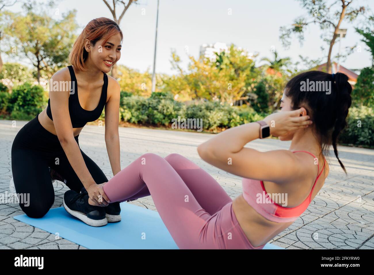 Sorridente giovane donna asiatica che regge le gambe di femmina amica seduta sul tappetino yoga che pratica crunches per abs al mattino in parcheggio Foto Stock