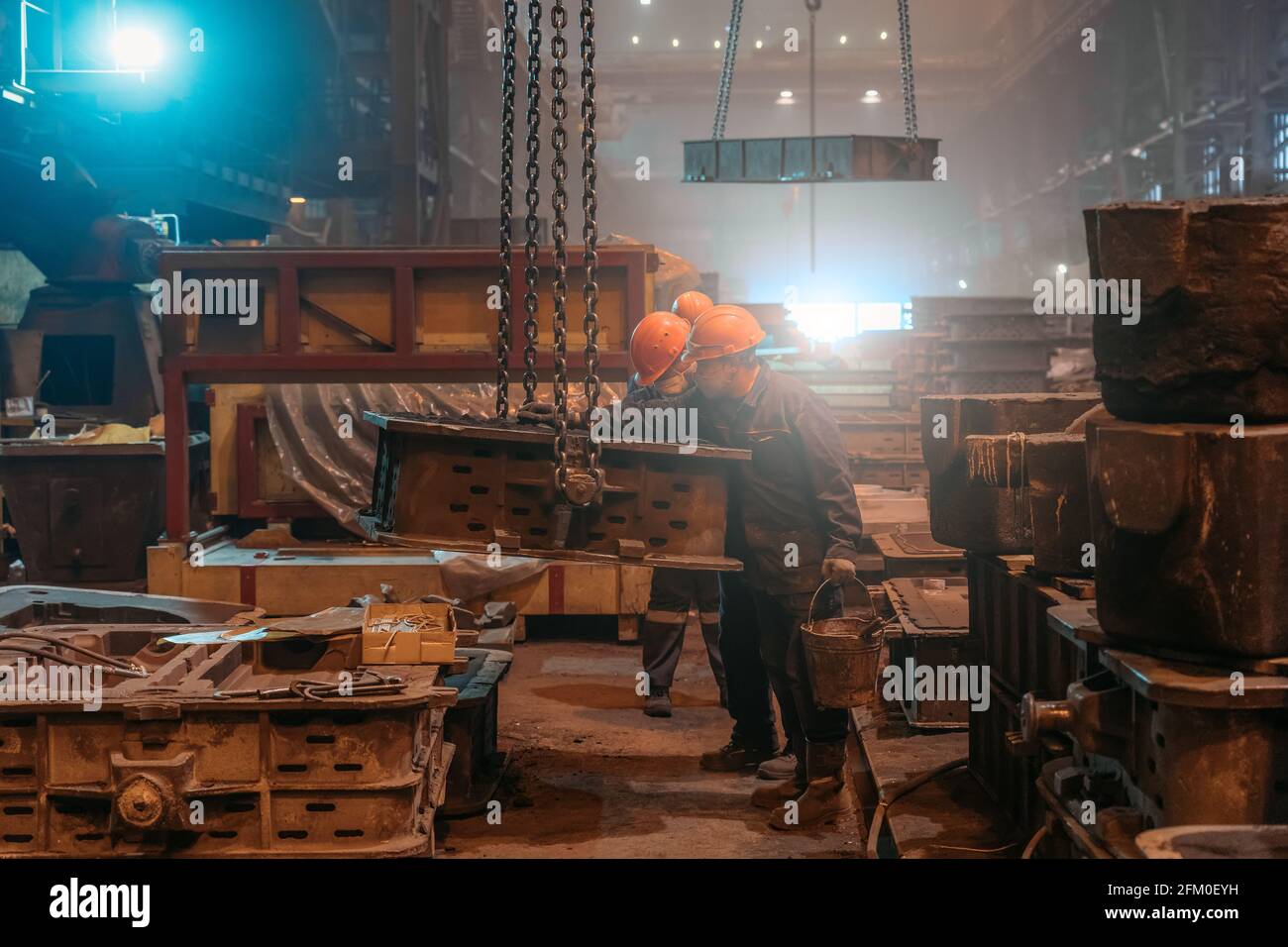 I lavoratori in caschi e maschere facciali lavorano con stampi su catene di gru dopo la fusione di metallo in fonderia di acciaieria. Foto Stock