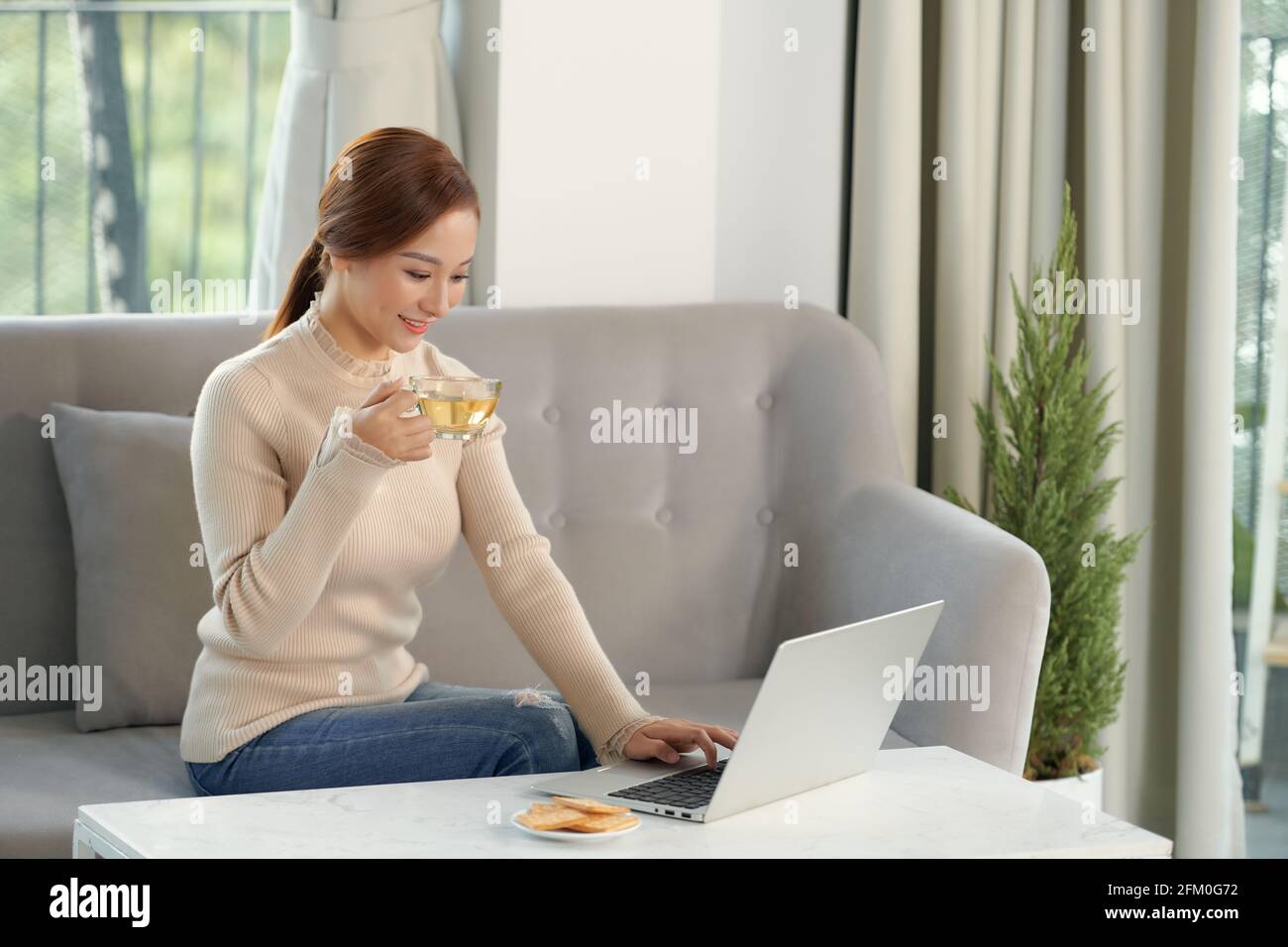 Bella ragazza allegra rossa che usa un computer portatile color argento mentre si siede sul divano in soggiorno a casa. Foto Stock