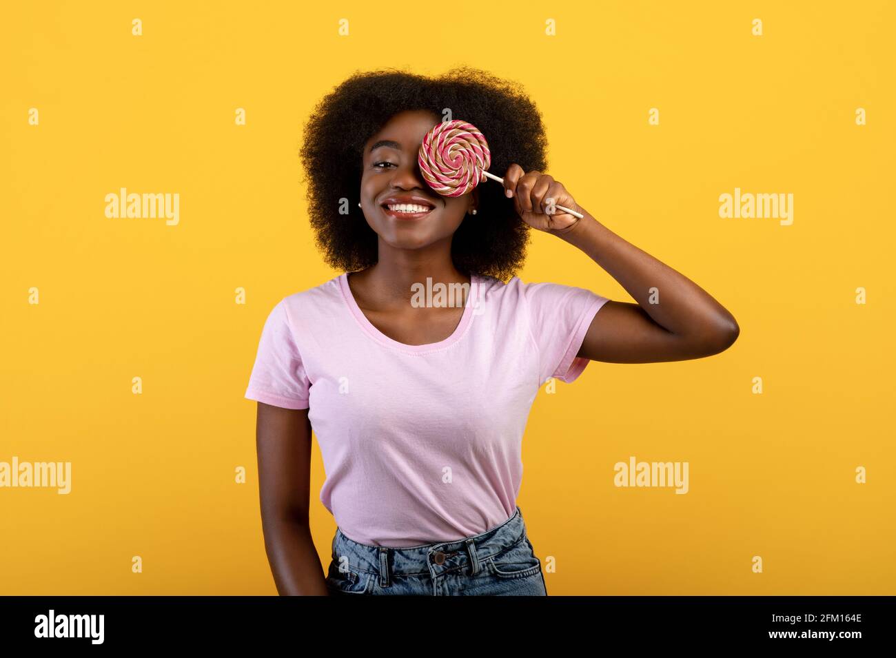 Dente dolce. Positiva giovane donna afroamericana che copre un occhio con lollipop su sfondo giallo luminoso studio Foto Stock