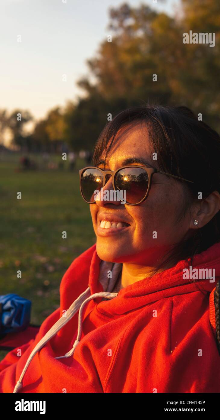 ritratto di giovane donna che indossa occhiali da sole in un parco verde loking al tramonto e sorridente Foto Stock