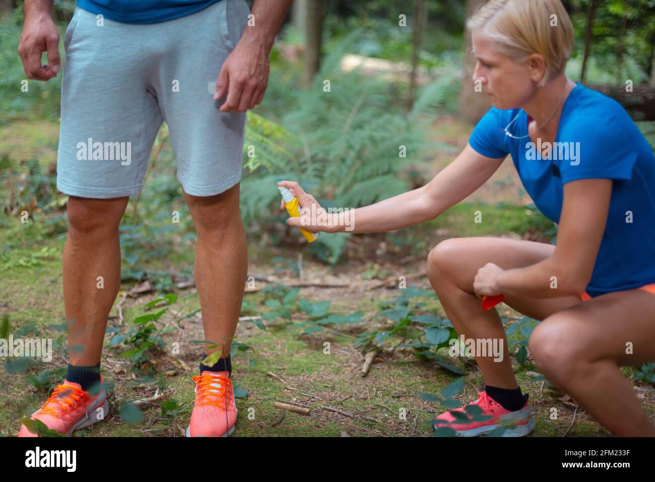 Un uomo in abbigliamento sportivo in piedi e una donna che si accovacciano accanto a lui, applicando zecche e zanzara repellente, spray, sulle gambe. Foto Stock