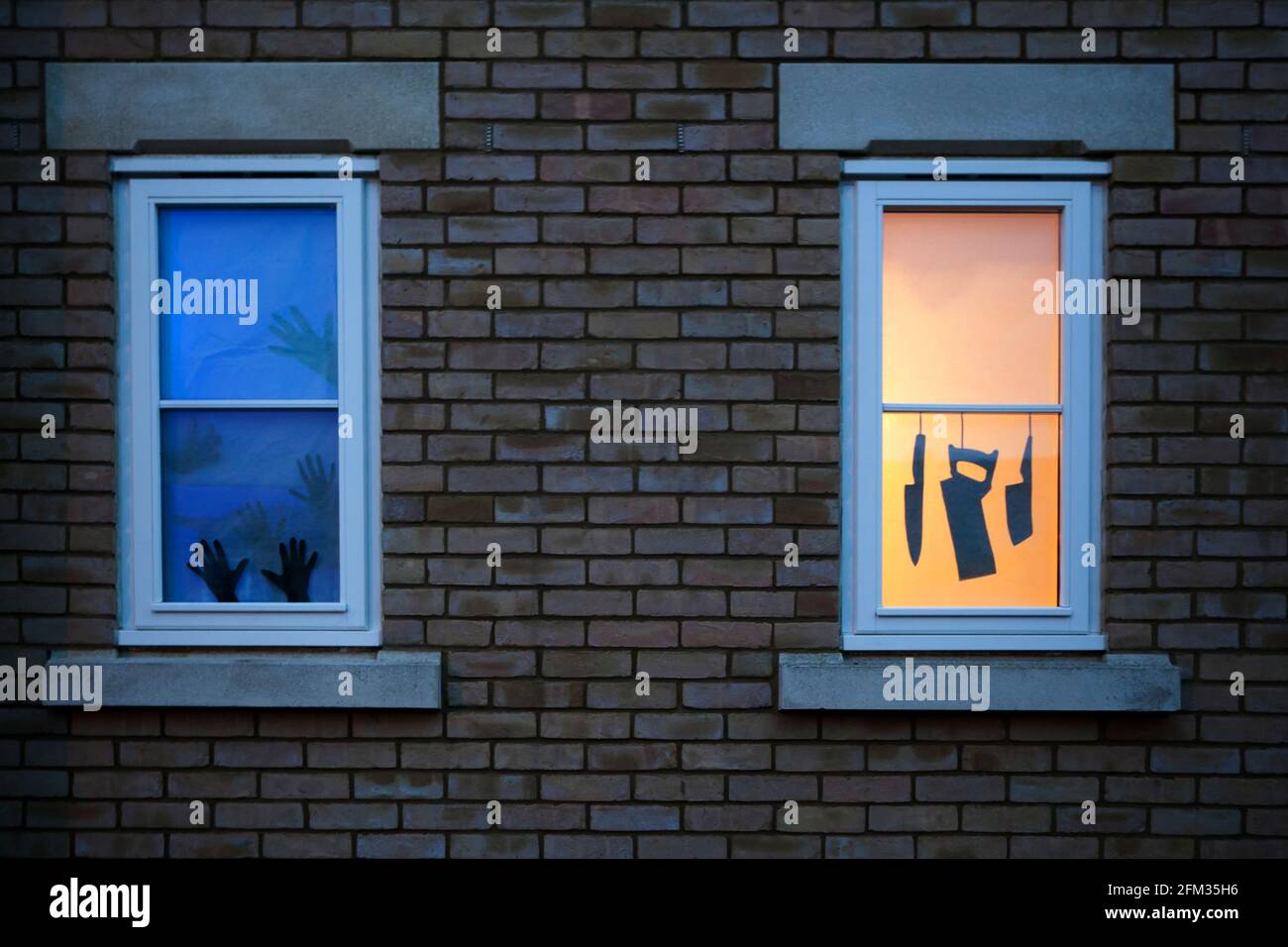Due silhouette di finestra a tema Halloween / casa infestata: Coltelli e una sega appesi in una finestra illuminata; mani che cercano di fuggire da un'altra. Foto Stock