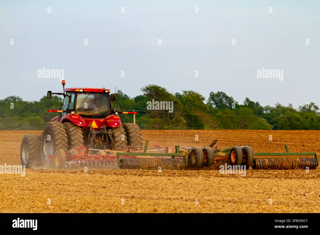 Un trattore a sei ruote rosso con cabina sta arando un campo agricolo vuoto nella molla. Lo strumento aratro collegato crea una nuvola di polvere nella fattoria. Imag Foto Stock