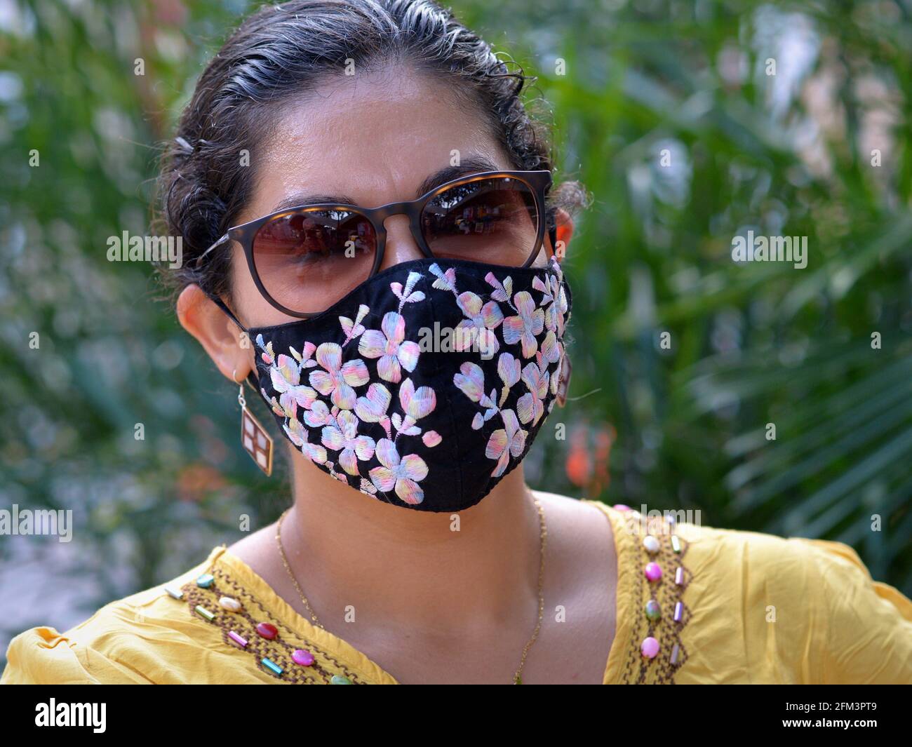 Bella giovane donna messicana affascinante con occhiali da sole marroni indossa una maschera elegante e posa per la fotocamera durante la pandemia di virus corona. Foto Stock