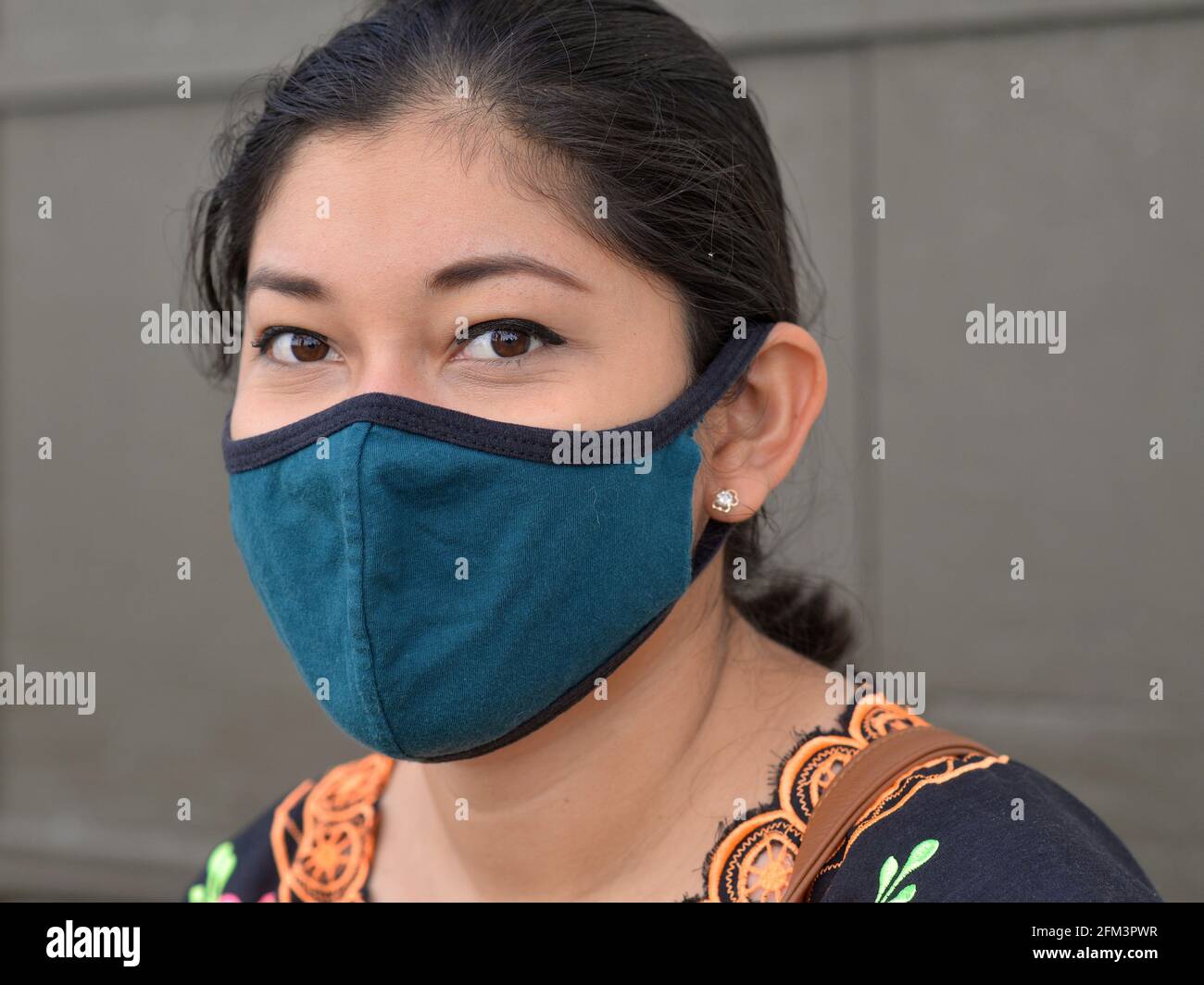 Affascinante giovane donna messicana dagli occhi marroni indossa una maschera di tessuto non medico blu durante la pandemia del coronavirus globale e guarda la fotocamera. Foto Stock