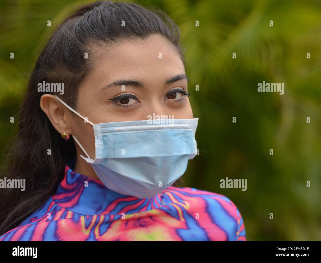Giovane bruna caucasica con gli occhi graziosi indossa una maschera chirurgica blu chiaro durante la pandemia globale del coronavirus e guarda lo spettatore. Foto Stock