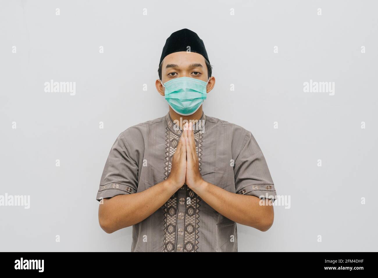 Un uomo musulmano indonesiano dà i saluti mettendo le mani di fronte al petto mentre indossa maschera medica, camicia musulmana e peci o songkok Foto Stock