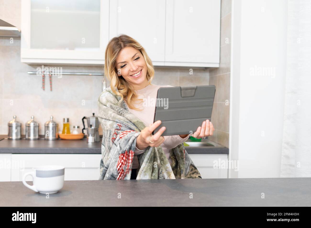 Bella donna sorridente gode di una videochiamata. Bionda ragazza a casa che tiene un tablet in cucina, seduta con le spalle coperte da una coperta calda. Foto Stock