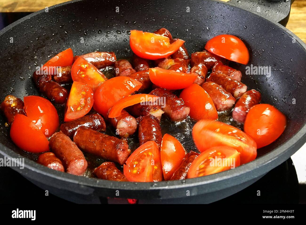 Deliziose salsicce di caccia alle grasse, tagliate a pezzi e il pomodoro a fette vengono fritte in padella in olio vegetale. Cibo nutriente veloce. Cucina domestica. Clos Foto Stock