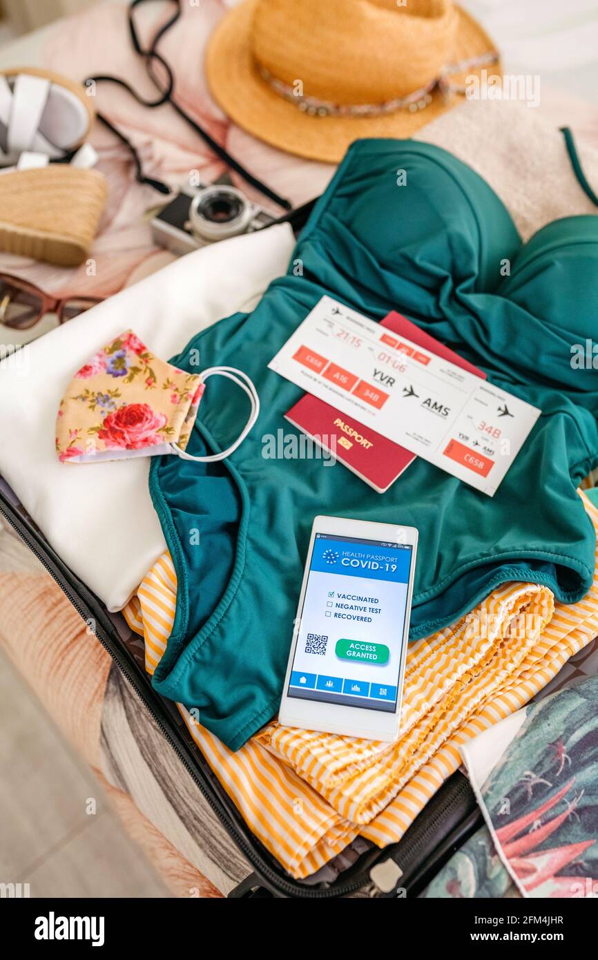 Vista dall'alto della valigia aperta con bagaglio da donna per la spiaggia passaporto delle vacanze e del covid sul telefono mobile Foto Stock