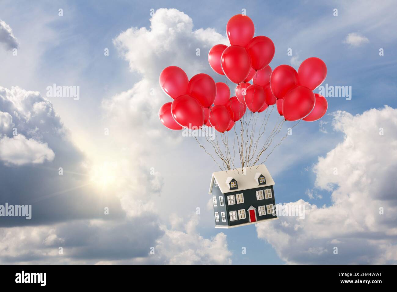 Casa giocattolo con palloncini rossi galleggianti nel cielo blu con sole e bianche nuvole soffici. Principi di casa in aumento e concetto di casa in movimento Foto Stock