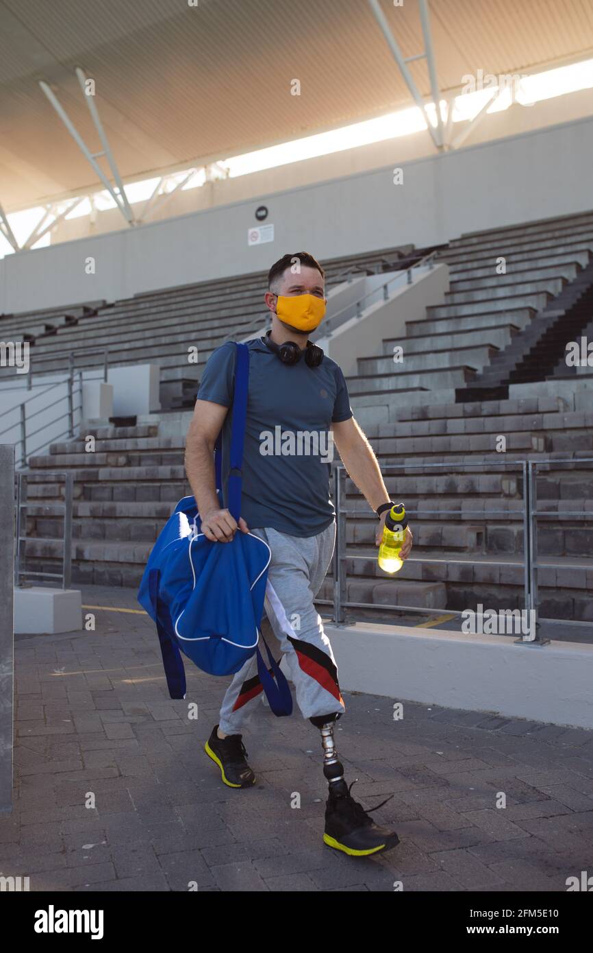 Atleta maschio caucasico con gamba protesica che indossa maschera facciale che cammina nello stadio Foto Stock