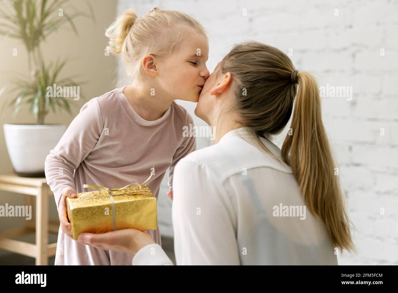 figlia piccola che dà il presente e il bacio alla sua mamma a casa. compleanno, giorno delle donne o concetto di giorno delle madri Foto Stock