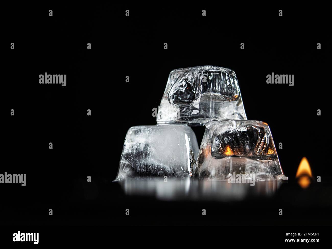 Tre cubetti di ghiaccio parzialmente fusi impilati su un riflettente nero Superficie e sfondo nero e due fiamme piccole nel Sfondo Foto Stock