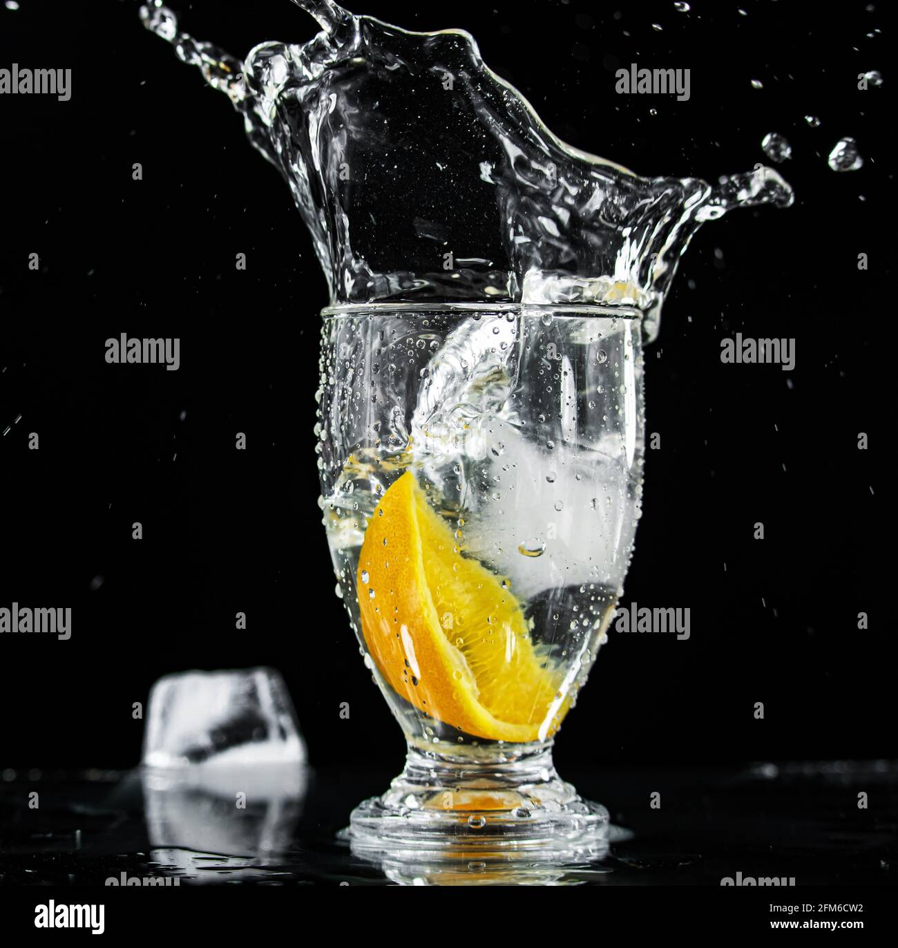 Vetro di acqua con la fetta arancione e goccia di ghiaccio che causa Acqua Splash su uno sfondo nero con un cubo di ghiaccio Sdraiato nelle vicinanze Foto Stock