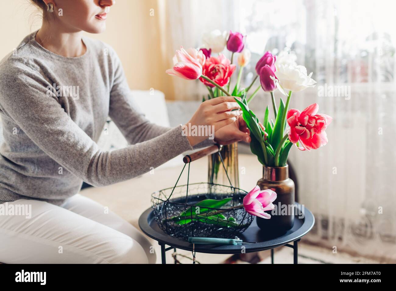 Donna organizza bouquet di fiori tulipani a casa utilizzando un cesto di metallo. Casalinga che si prende cura di intimità in appartamento. Interni e decorazioni Foto Stock
