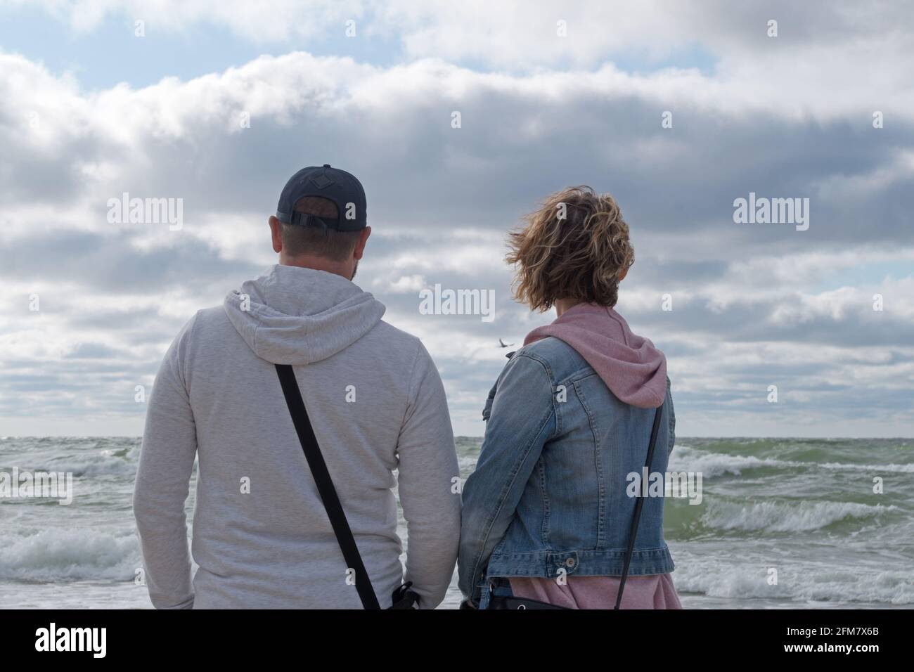 Un paio di giovani, un uomo in giacca grigia e una donna in giacca blu denim si levano di fronte al mare in una giornata ventosa sullo sfondo di una tempesta Foto Stock