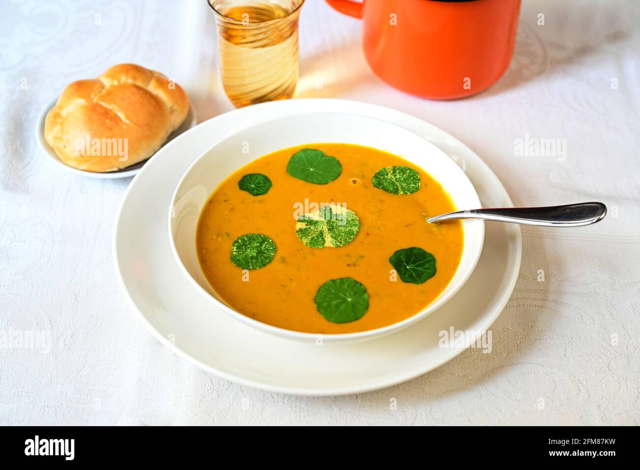Zuppa di zucca con foglia verde commestibile (nasturzio) in piatto bianco, pane, bicchiere d'acqua e vaso d'arancia su tavola bianca. Foto Stock