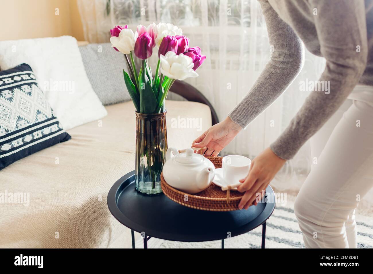 Donna mette il bollitore e la tazza del tè sul vassoio di rattan sul tavolino da caffè. Interni e arredamento accogliente. Tulipani freschi fiori messi in vaso Foto Stock