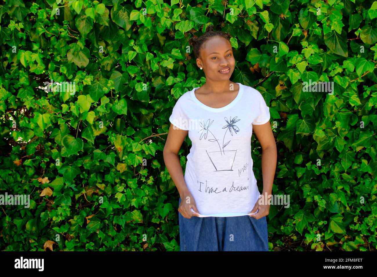 Giovane donna africana con uno stile fresco capelli e una t-shirt in posa all'aperto in un giardino con uno sfondo foglie. Foto Stock