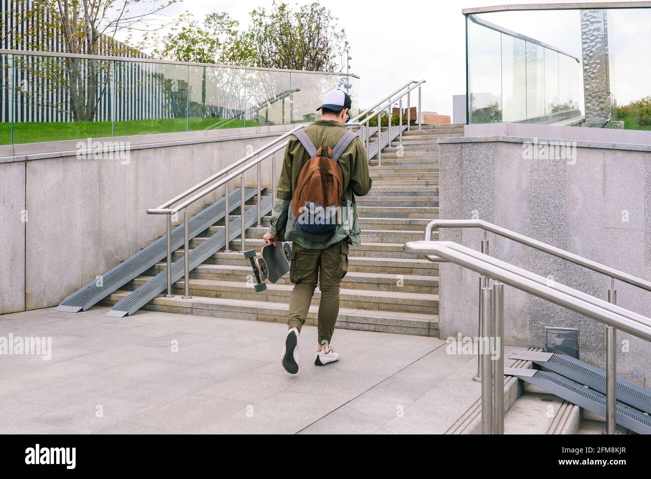 Il giovane cammina per la strada con uno skateboard e ascolta la musica con le cuffie. Giovani attivi in un ambiente urbano. Vista posteriore Foto Stock