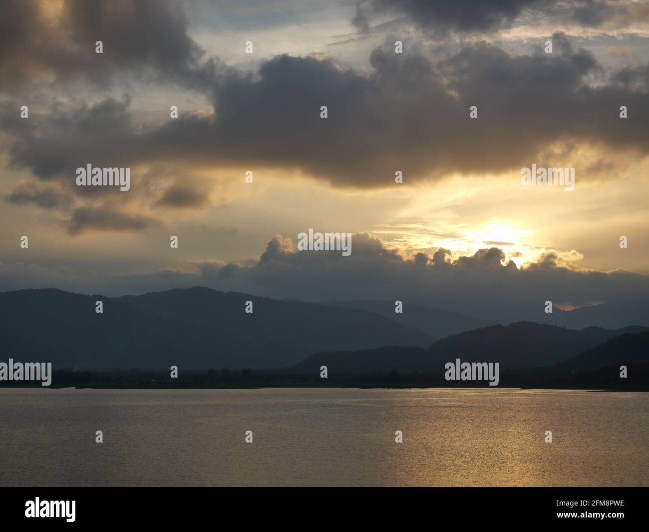 Il sole giallo dietro la sagoma della nuvola e della montagna si riflette sulla superficie del lago al tramonto, la superficie dell'acqua scintilla Foto Stock