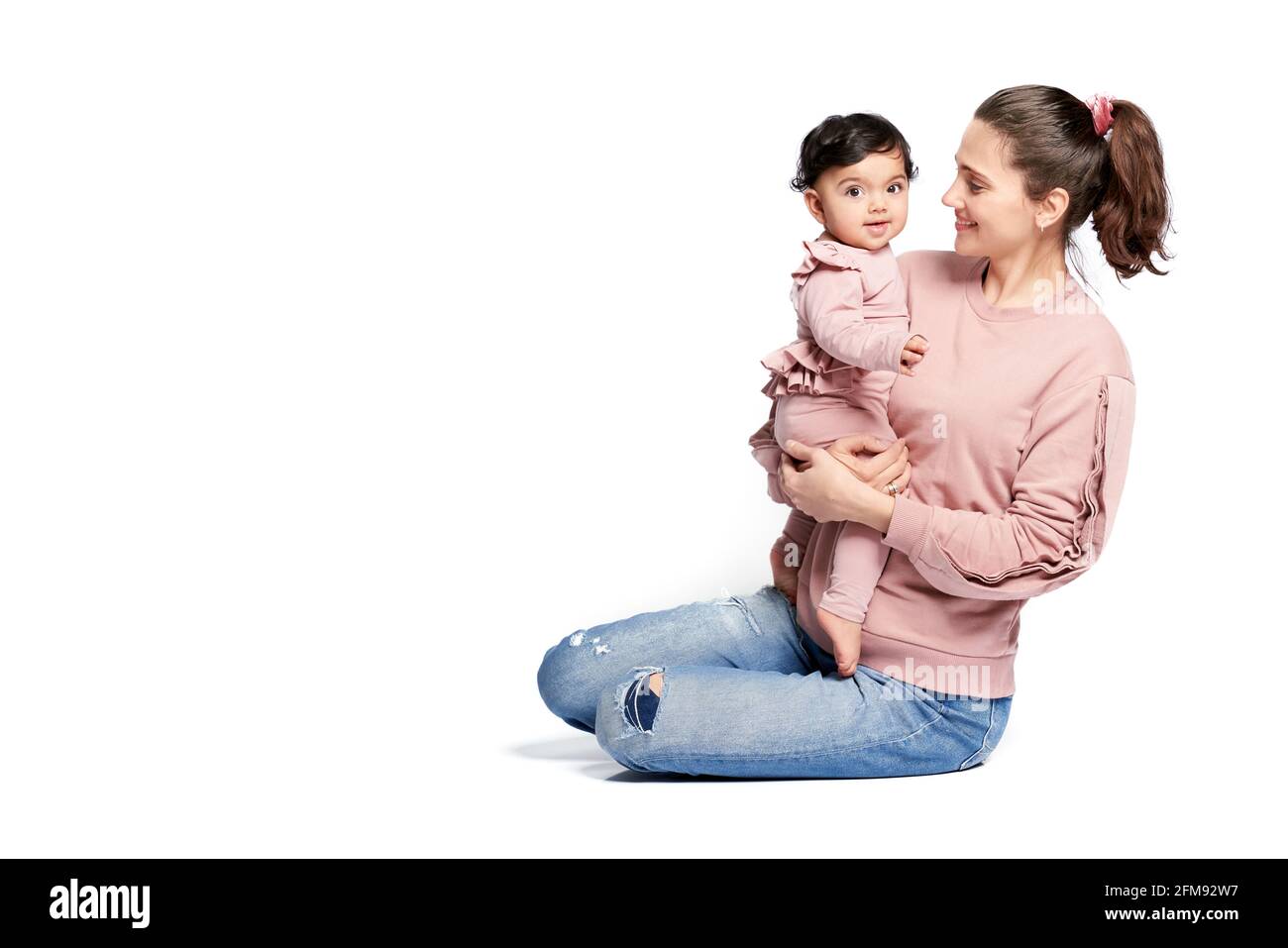 Ritratto di madre con figlia sorridente isolato su sfondo bianco studio. Vista laterale della giovane donna che tiene il bambino adorabile dolce in braccio mentre si siede sul pavimento, bambino felice guardando la macchina fotografica. Foto Stock