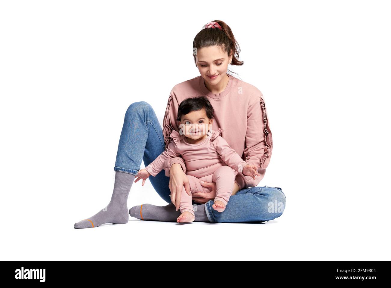 Ritratto di madre carina con figlia sorridente isolato su sfondo bianco studio. Giovane donna attraente che tiene il bambino adorabile dolce sulla gamba mentre si siede sul pavimento, concetto felice di infanzia. Foto Stock