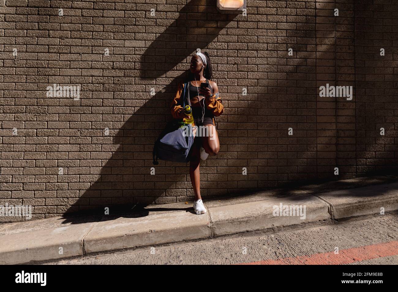Fit donna afroamericana con borsa da palestra e bottiglia d'acqua appoggiandosi al muro di mattoni in città Foto Stock