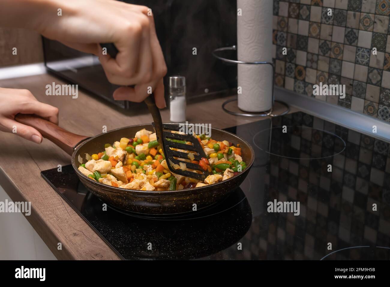 Le mani delle donne si mescolano con una spatola a pezzi di carne bianca, una varietà di verdure che vengono fritte in una padella su una stufa a induzione primo piano. Foto Stock