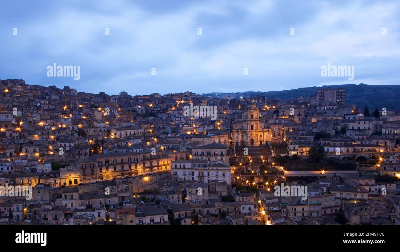 Italia, Sicilia, sud-est, angolo barocco, Modica, Il crepuscolo, la vista di Modiac e del Duomo di San Giorgio, la cattedrale e le case del centro storico sono già illuminate di luce artificiale, il cielo sopra è grigio-blu e nuvoloso Foto Stock