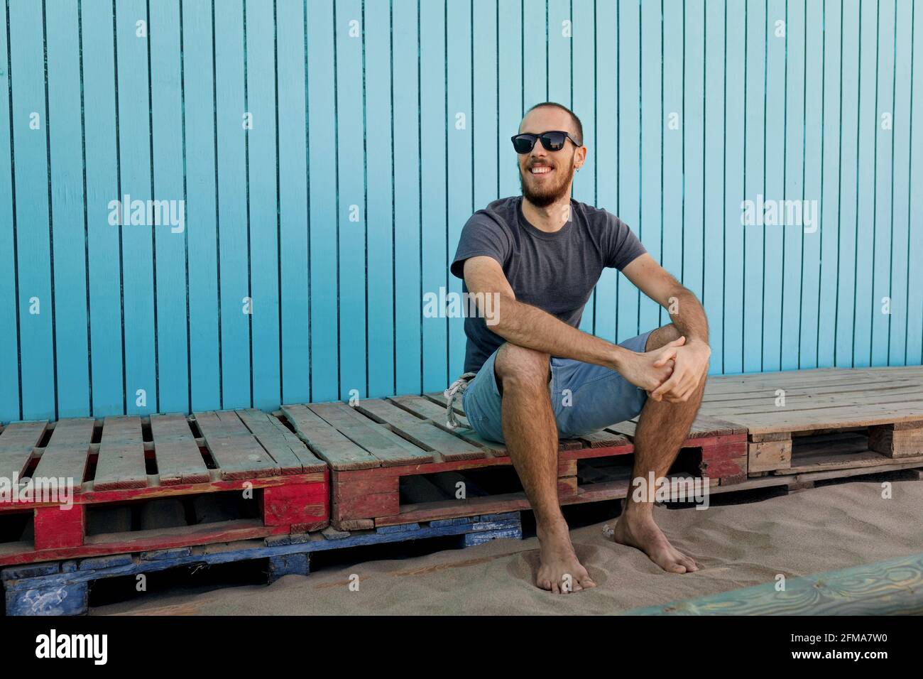 Felice giovane bearded indossare occhiali da sole sta sorridendo a Isla Canela spiaggia seduta di fronte ad una parete blu di legno mentre guardando via Foto Stock