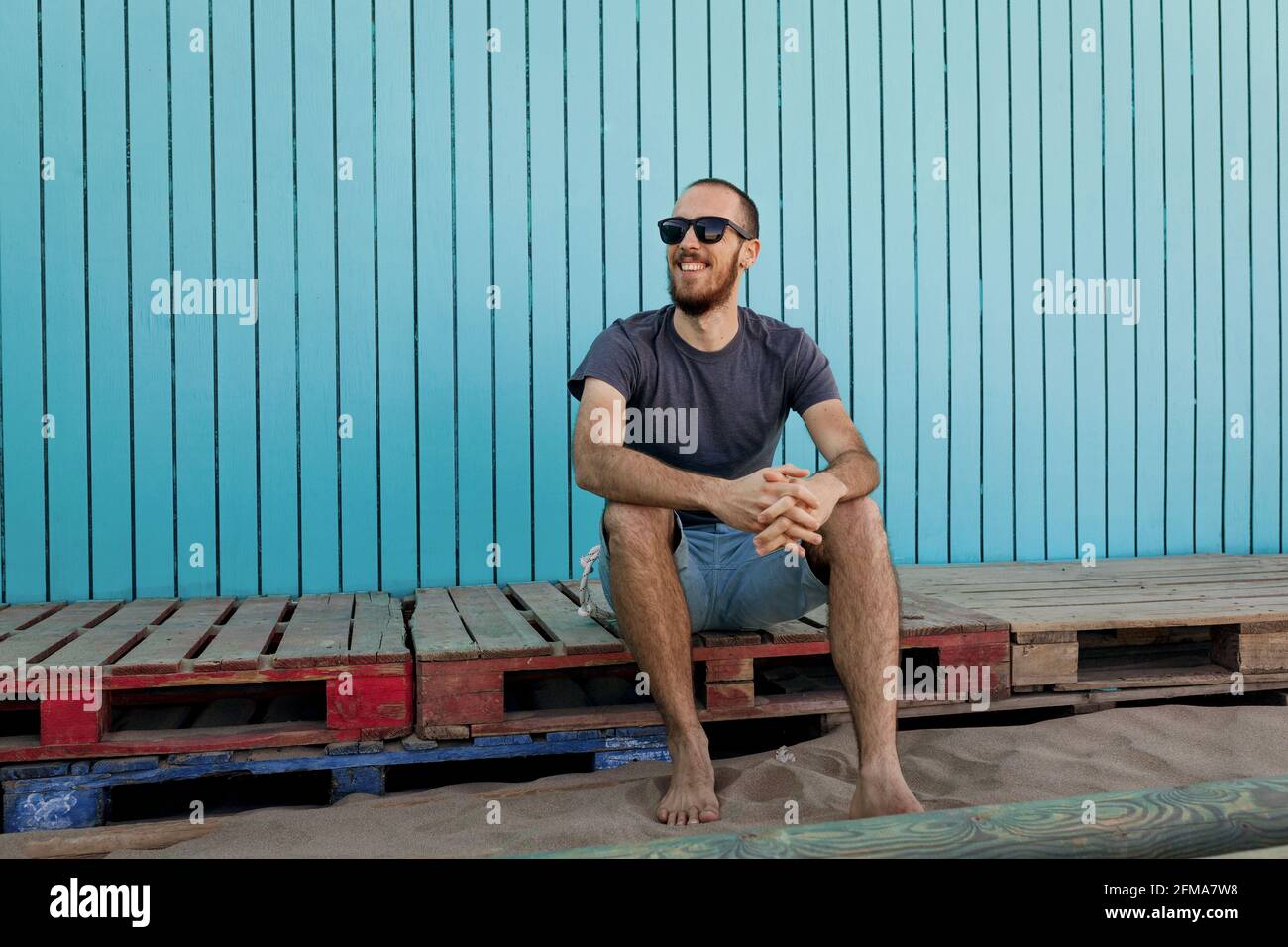 Felice giovane bearded indossare occhiali da sole sta sorridendo a Isla Canela spiaggia seduta di fronte ad una parete blu di legno mentre guardando via Foto Stock
