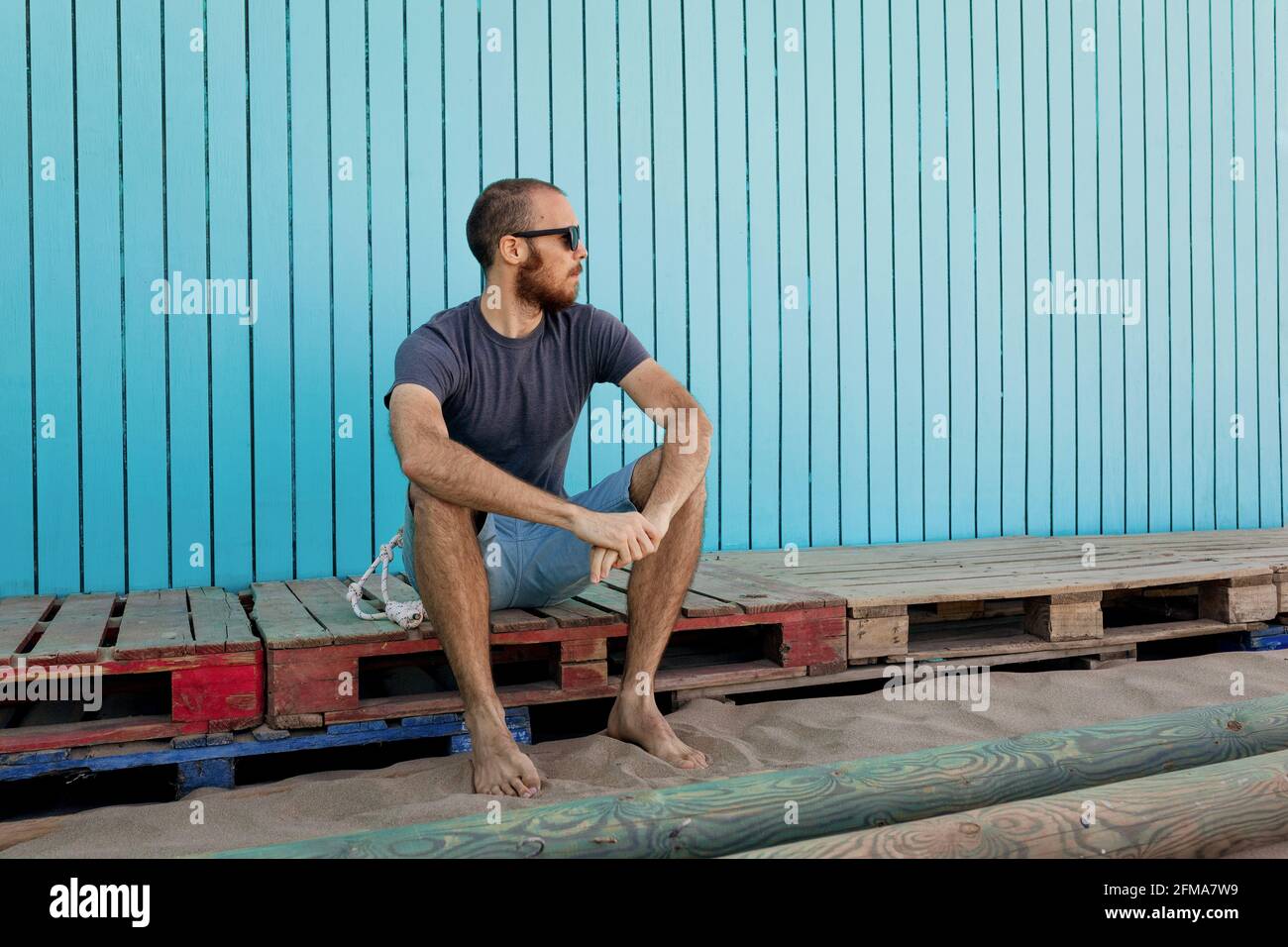 Il giovane uomo bearded che indossa gli occhiali da sole sta guardando via sicuro La spiaggia di Isla Canela con pareti blu in legno Foto Stock