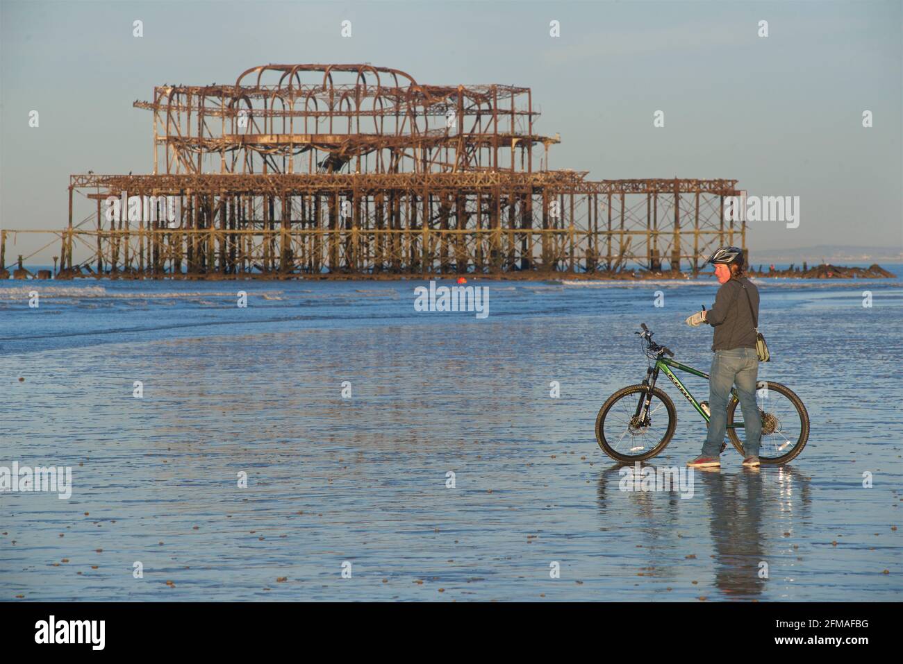 Ciclista in piedi con mountain bike sulla sabbia a bassa marea, Brighton, con i resti arrugginiti del West Pier sullo sfondo. Brighton & Hove, Sussex, Inghilterra Foto Stock