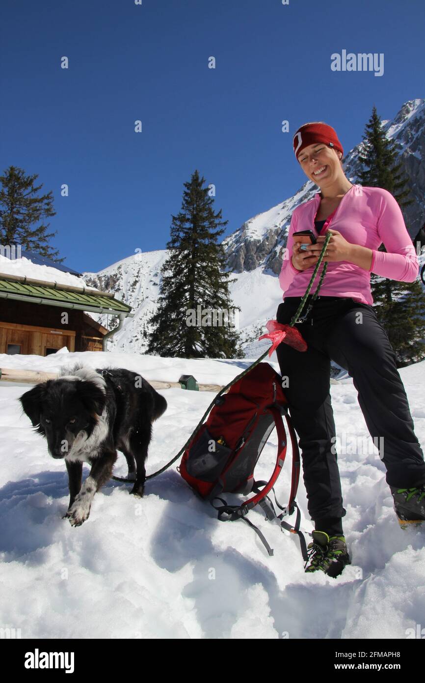 giovane donna con cane e telefono cellulare di fronte alla capanna altopiano, 1623 m, chiuso, riposo invernale, ristorante, Escursione invernale nei pressi di Mittenwald, sullo sfondo i monti Wörner, Wörnersattel, Karwendel, Europa, Germania, Baviera, alta Baviera, Valle d'Isar, Werdenfelser Land, altopiano, DAV, Rifugio Club Alpino, Club Alpino Tedesco Foto Stock